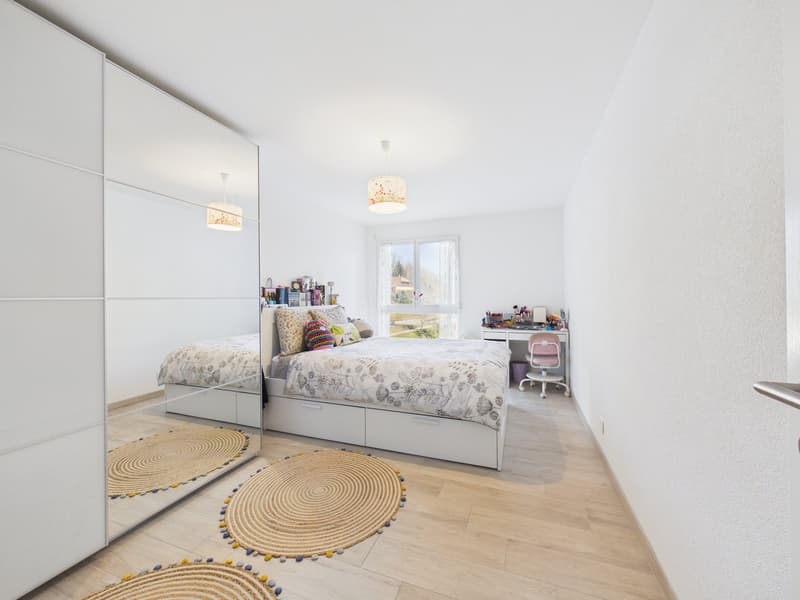 A clean white bedroom with a double bed, a mirror on the wall, two lamps, two round rugs, and a desk with a chair.