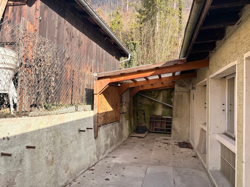 Old building with wooden walls, concrete floor, metal fence, wooden pergola, and a small shelf in the corner.