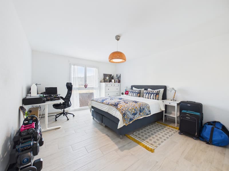 Bedroom with bed, desk, chair, luggage, gym equipment, rug, lamp, cabinet, window, and various items on the desk and cabinet.