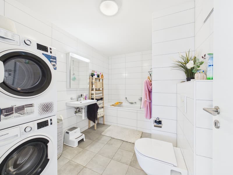 Two stacked washing machines, mirror above sink, bathtub, tiled floor, showerhead, plant, towel rack