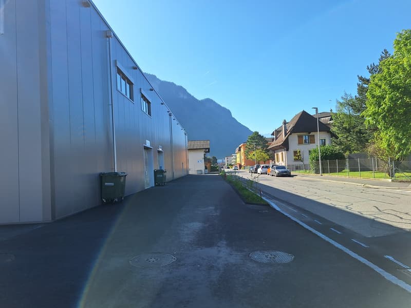 Industrial building, gray metal siding, windows, dumpsters, empty road, parked cars, houses, trees, mountain in background, clear sky.