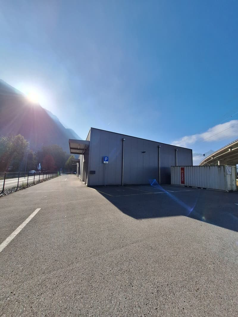 A large commercial building with a parking lot in front. The building has a simple, modern design with a dark exterior. The parking lot is paved and has a designated parking space. The surrounding landscape includes mountains and a bright, sunny sky.