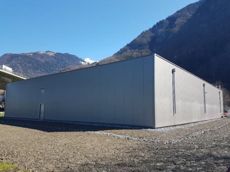 A large, rectangular industrial or commercial building with a flat roof and gray exterior walls. The building is situated in a mountainous area with a clear blue sky in the background.