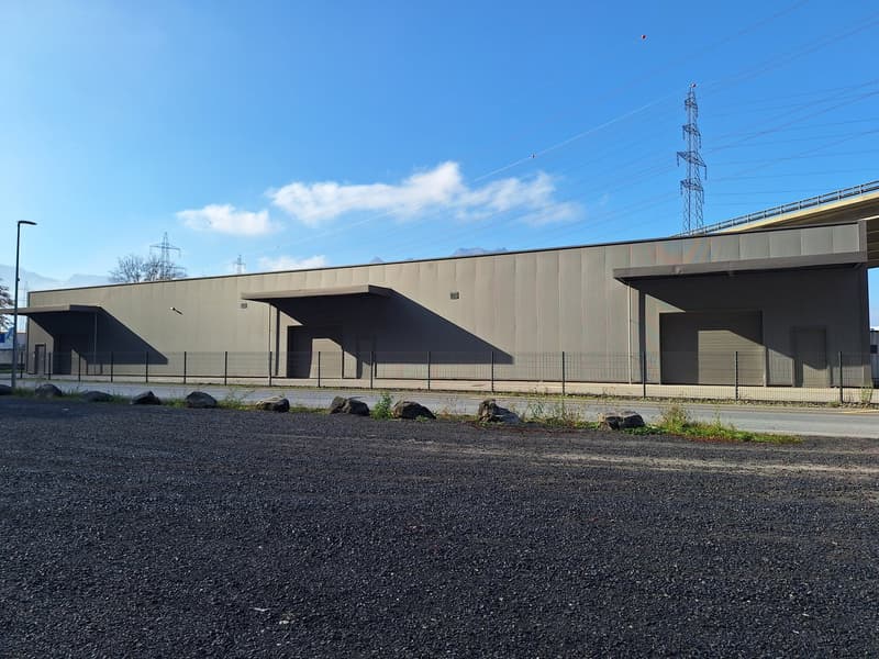 A large industrial or commercial building with a flat roof and multiple entrances. The building has a gray and black exterior with a paved parking area in front.