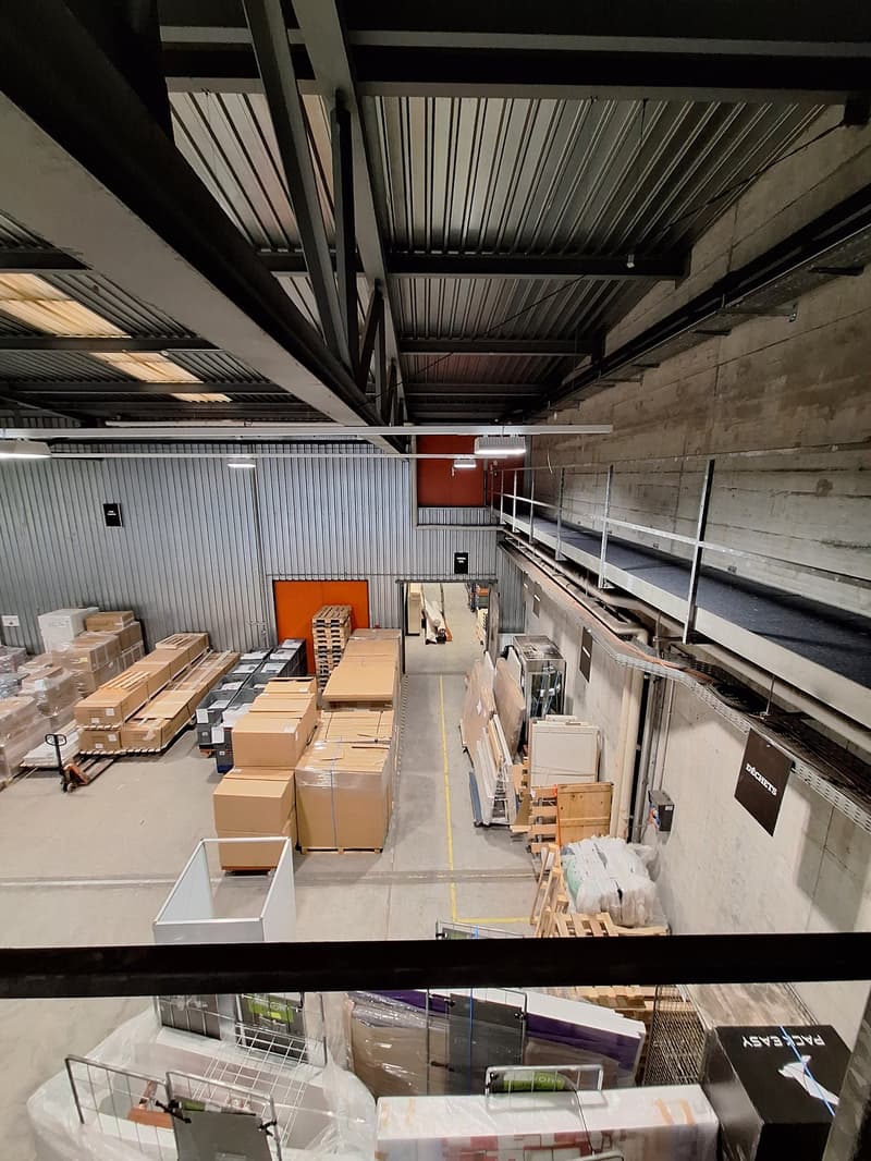 large warehouse with high ceilings, metal beams, concrete walls, multiple storage boxes, wooden pallets, metal railings, and a mezzanine