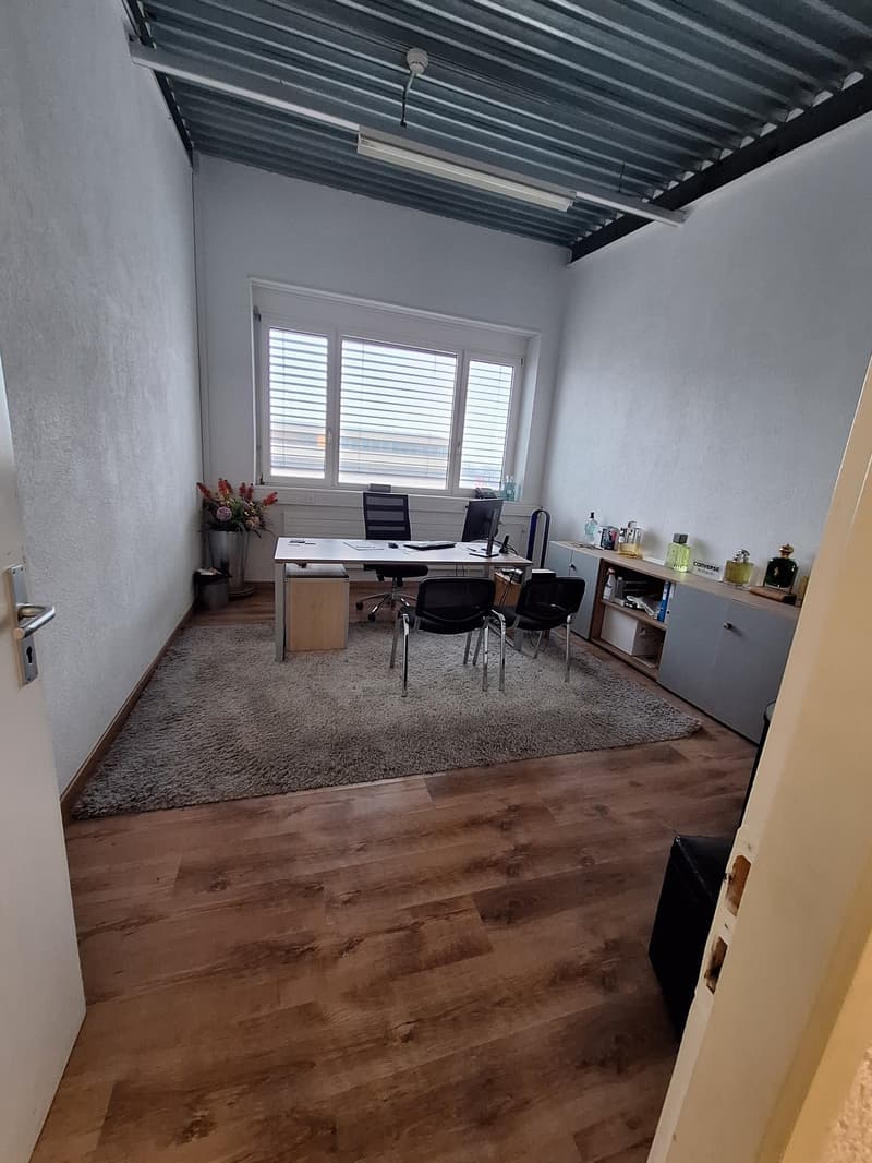 empty office room with wooden floor, white walls, desk, chairs, window with blinds, rug, cabinet, trash bin, flower vase