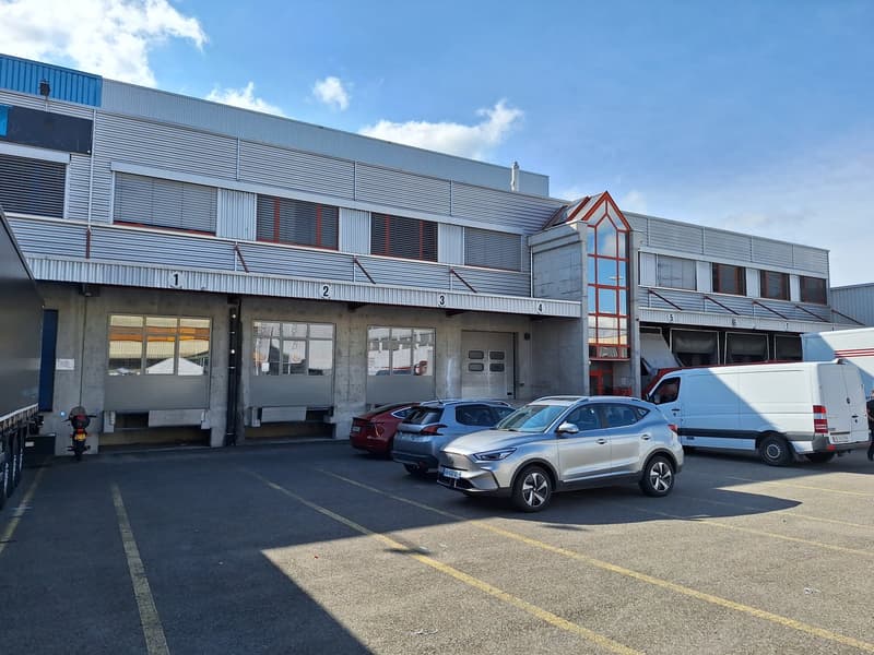 Industrial building with multiple windows, large garage doors, and a parking lot with several cars parked.