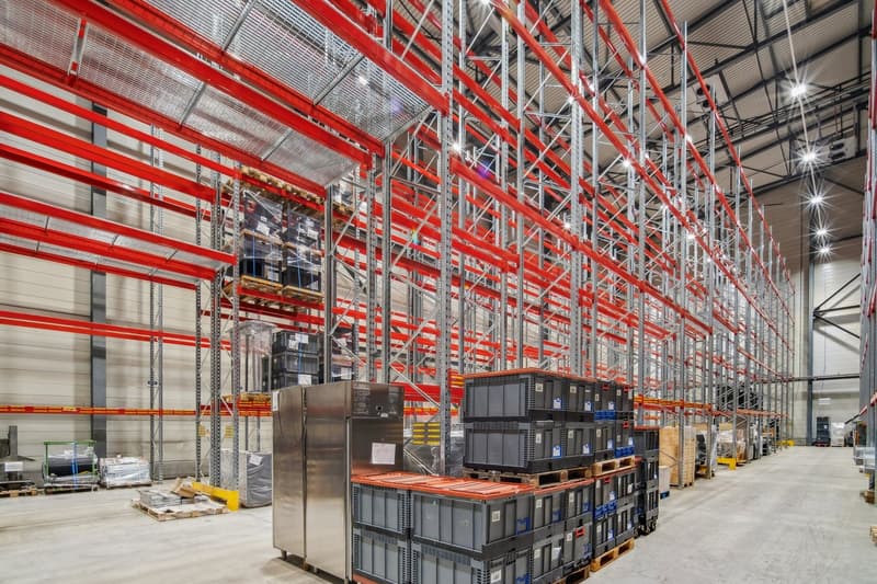 large warehouse with red and silver metal shelving, multiple stacked boxes, large silver refrigerator, concrete floor