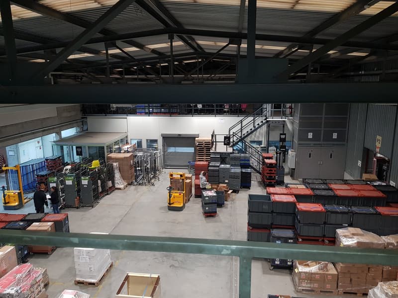large warehouse with multiple pallets, forklifts, and storage boxes, mezzanine level, workers present