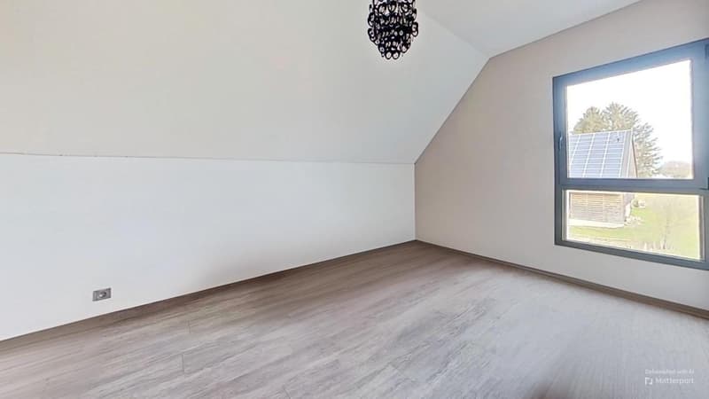 Spacious bedroom with white walls, hardwood-style flooring, and a large window providing natural light. The room features a modern black chandelier as the main lighting fixture.