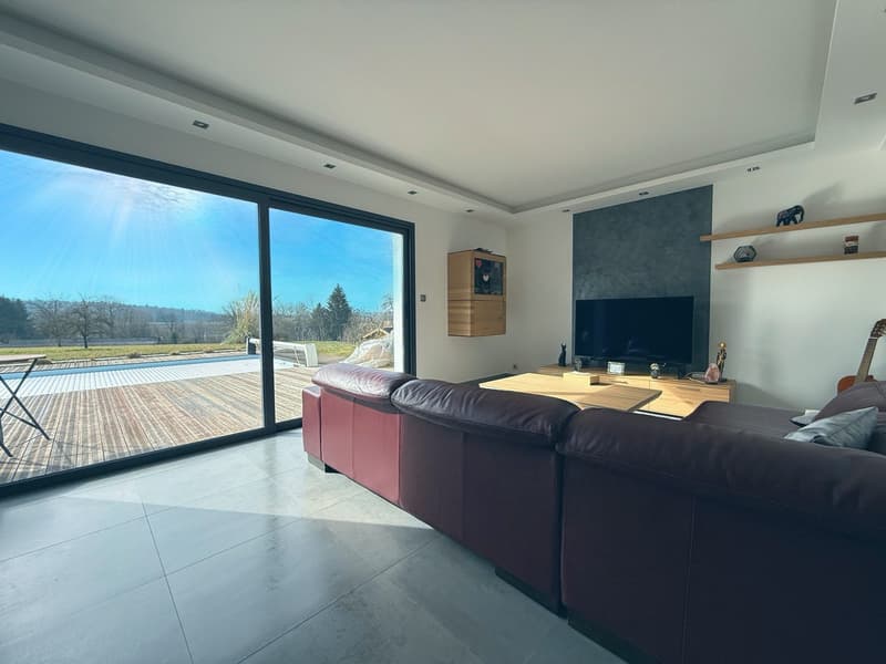 Spacious living room with large windows overlooking an outdoor pool and scenic landscape, dark leather sofa, wooden shelves, and a TV mounted on the wall