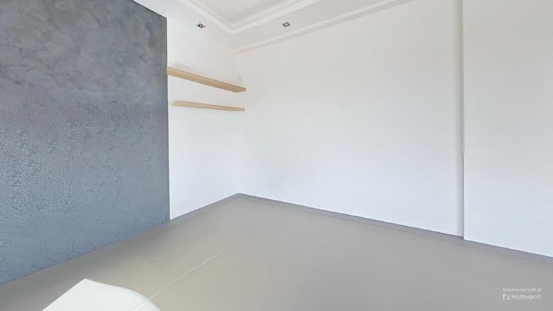 The image shows a minimalist room with white walls, a gray accent wall, and wooden shelves. The floor appears to be a light-colored laminate or tile. The room has a clean, modern aesthetic with a simple and uncluttered design.