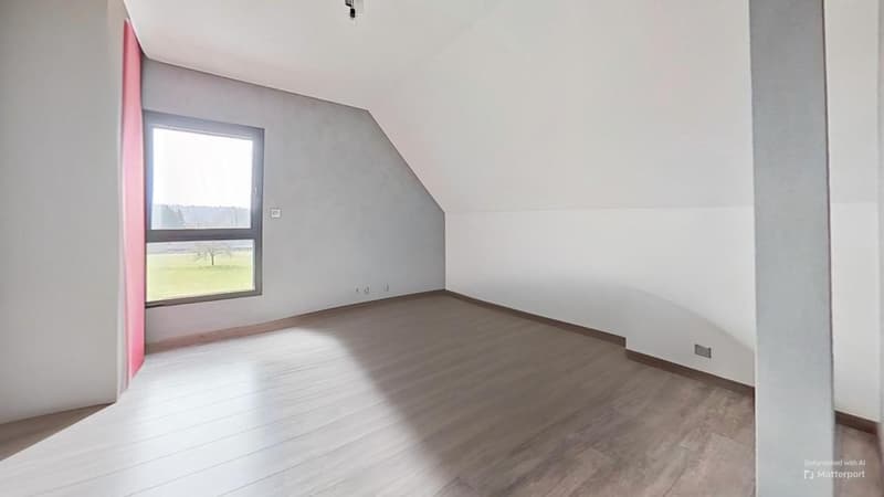 Spacious bedroom with slanted ceiling, large window providing natural light, hardwood-style flooring, and a minimalist design with white walls.