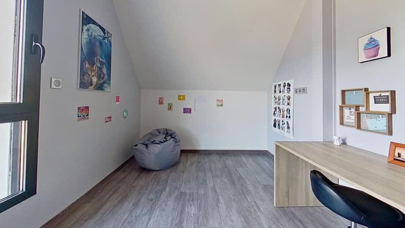 Spacious bedroom with hardwood flooring, white walls, and various decorative elements such as posters and framed artwork. A large bean bag chair is placed in the corner, and a desk with a chair is situated against the wall.