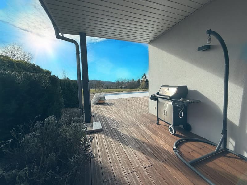 Covered outdoor patio with wooden deck, gas grill, and a view of the swimming pool and surrounding landscape.
