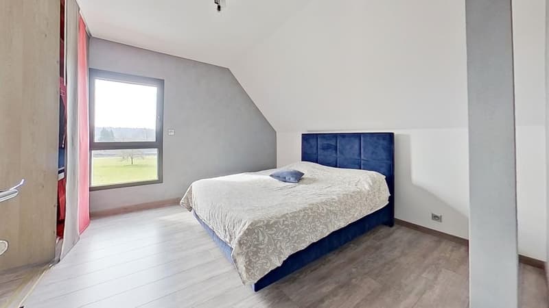 Spacious bedroom with large window providing a view, grey walls, hardwood flooring, and a blue upholstered bed frame with a patterned comforter