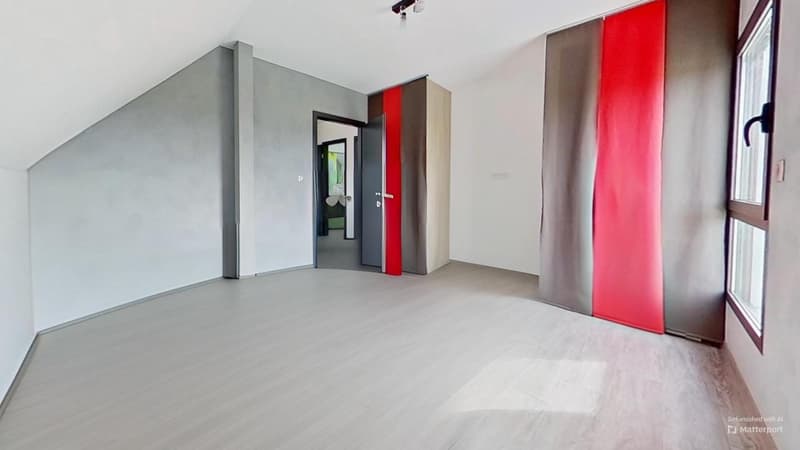 Spacious bedroom with gray walls, hardwood flooring, large window, and red curtains. The room has built-in closets and a doorway leading to another room.
