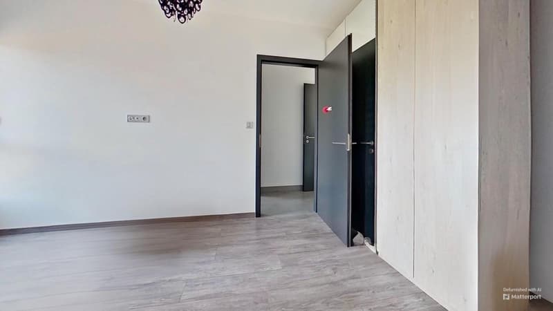 The image shows an empty room with white walls, a hardwood-style floor, and a black door frame. There is an electrical outlet visible on the wall. The room appears to be a living space or bedroom.