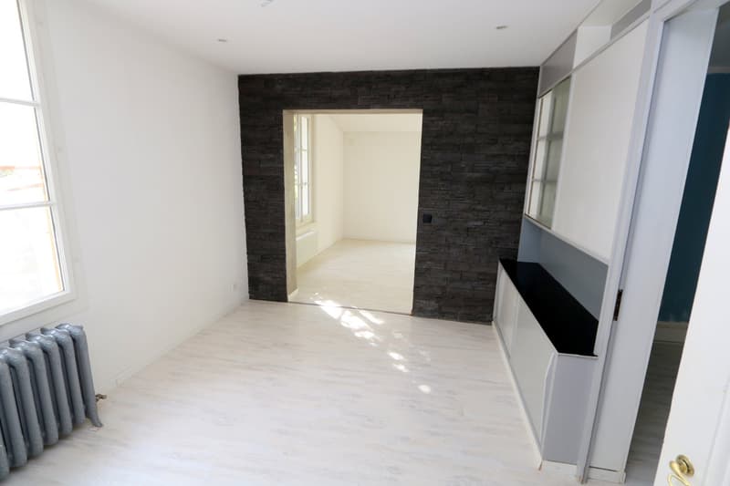 empty room with white walls, radiator, wooden floor, door leading to another room with a window