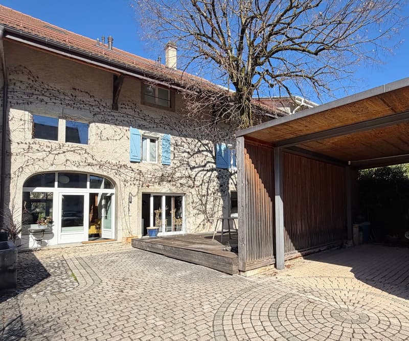 stone house with multiple windows, brick walls, chimney, wooden garage with canopy, brick paved courtyard, and a tree