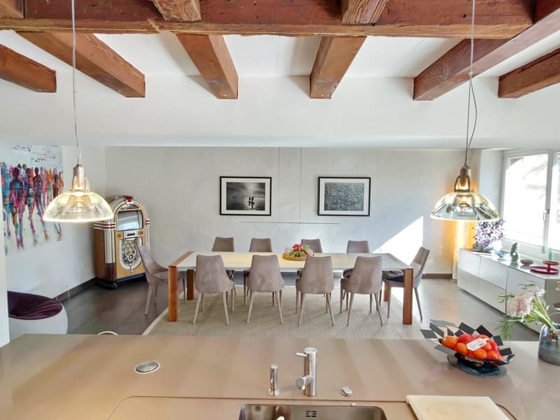 spacious room with wooden beams, white walls, central dining table with chairs, modern kitchen with sink and faucet, carpeted floor, decorative items, paintings, vintage jukebox, and a window with a view