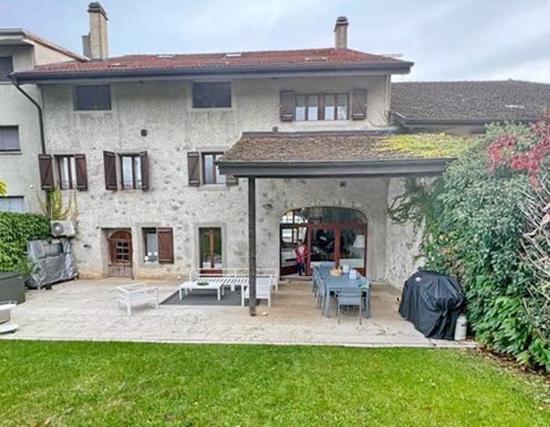 Stone-walled house with a covered patio and garden. Features include red tiled roof, brown wooden shutters, and an outdoor seating area with tables and chairs.