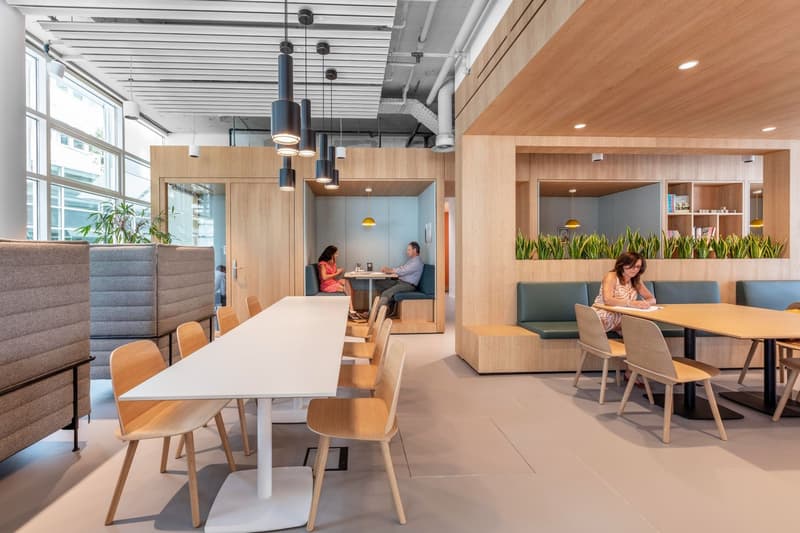 An open office space with several desks, chairs, and plants. A long white desk with four chairs is at the front, and a large green sofa is to the right.