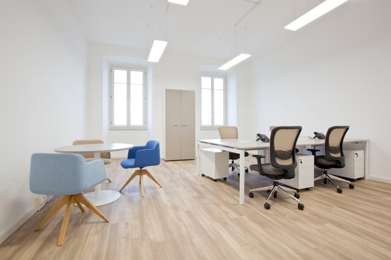Open-space office, wooden floor, white walls, white desk, blue and beige office chairs, round table, storage cabinet, windows