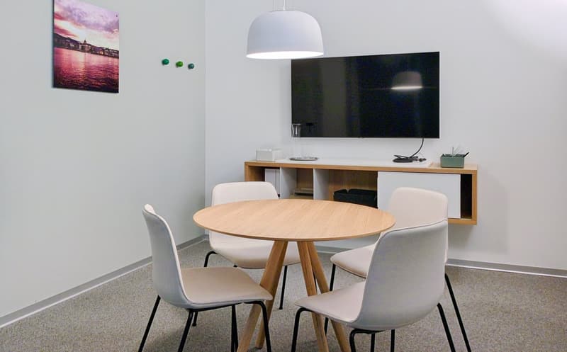 The image shows a modern living room with a round wooden dining table and four white chairs. There is a large TV mounted on the wall above a wooden media console. The room has white walls and a gray tile floor. A large pendant light fixture hangs over the