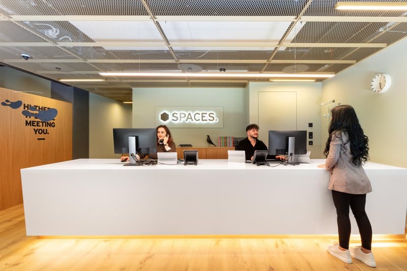 Open office space with reception desk, computers, and employees working at desks. The space has a modern, minimalist design with wooden accents and a large 'SPACES' logo on the wall.