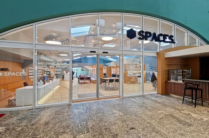 Large open office space with glass walls, wooden accents, and modern furniture. The space is labeled 'SPACES' and has a welcoming message on the glass. There appears to be a parking area or entrance in front of the office.