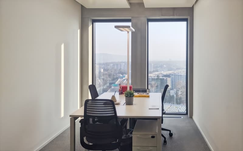 Spacious office with large windows offering a panoramic view of the city skyline. The room features a long wooden desk with two black office chairs, a potted plant, and some office supplies. The lighting is bright and the overall atmosphere appears modern