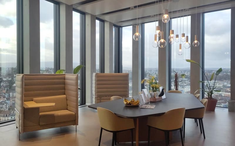 Spacious living room with floor-to-ceiling windows offering a panoramic view of the city. The room features a large dining table with comfortable chairs, and a cozy seating area with plush armchairs. The space is illuminated by a collection of modern pend