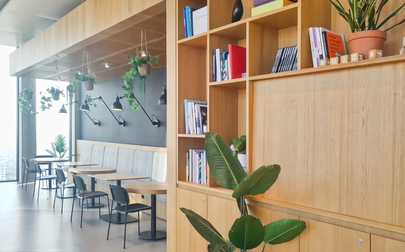 Modern cafe interior with wooden shelving, potted plants, and a seating area with tables and chairs. The space features large windows, pendant lighting, and a bookshelf filled with books and decor items.
