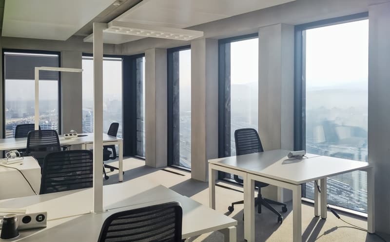 Spacious office with large windows offering a panoramic view of the city skyline. The office features modern, minimalist furniture including desks and chairs. The lighting is bright and the overall atmosphere is professional and conducive to work.