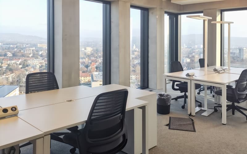 Spacious office with large windows offering a panoramic view of the city skyline. The office features multiple workstations with desks and ergonomic office chairs. The decor is minimalist with a neutral color scheme and modern lighting fixtures.