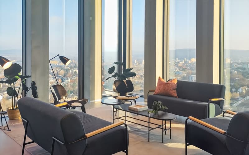 Spacious living room with floor-to-ceiling windows offering a panoramic view of the city skyline. The room features modern, minimalist furniture including black leather armchairs, a black sofa, and a glass coffee table. Decorative elements like plants and