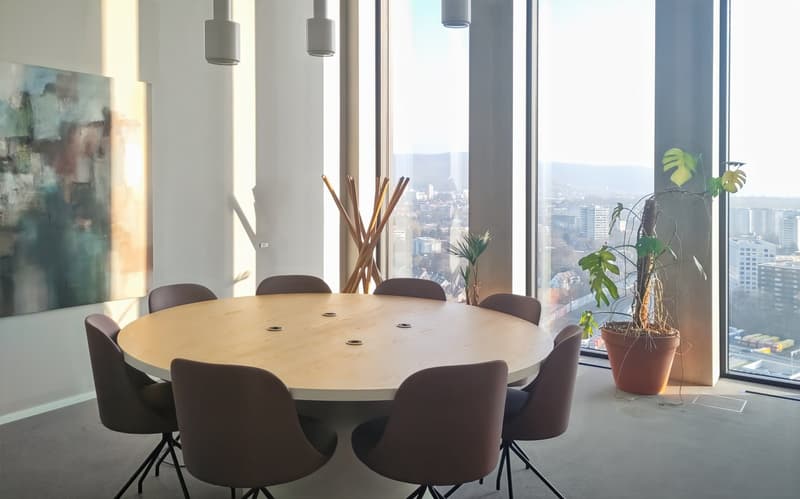 Large round wooden dining table with 8 brown leather chairs, modern pendant lighting, abstract art on the wall, and a large window with a city view