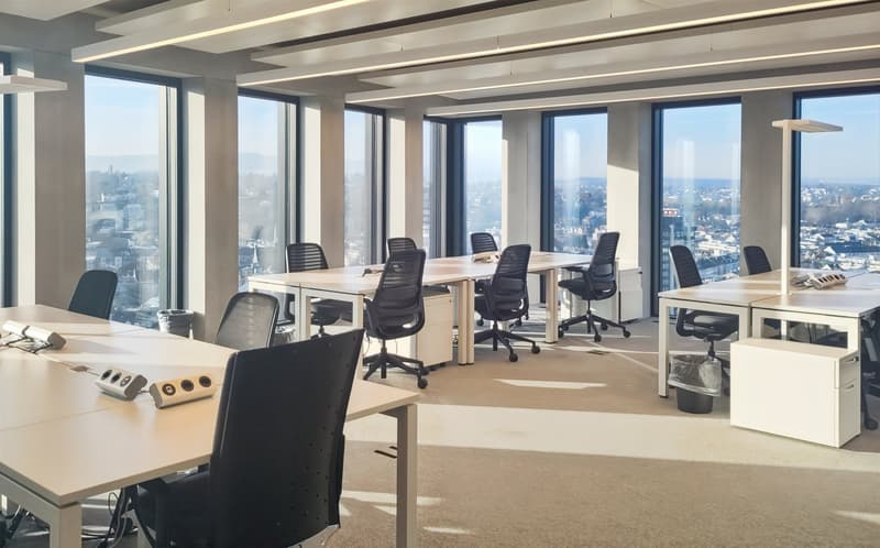 Large open-plan office space with floor-to-ceiling windows offering a panoramic view of the city skyline. The office features multiple workstations with desks and ergonomic office chairs, as well as various electronic devices and equipment. The space has