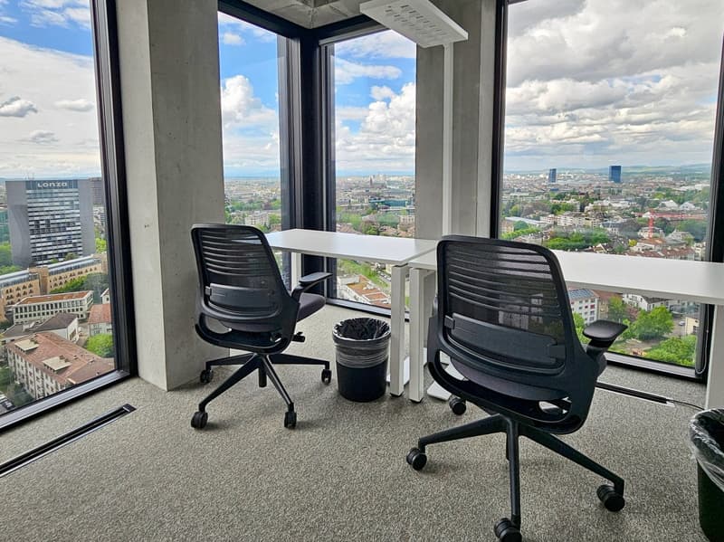 High-rise office space with large windows offering a panoramic view of the city skyline. The space features a small table and two black swivel chairs, as well as a potted plant. The office appears to be located on a high floor, providing an elevated persp