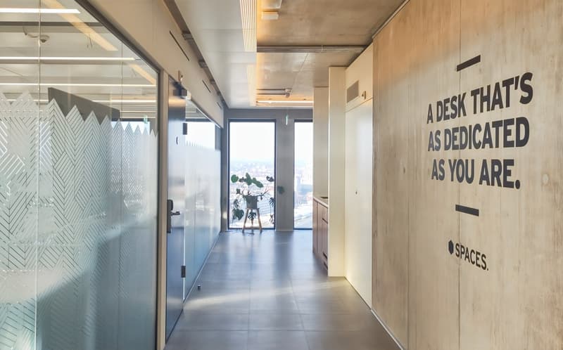 Modern office space with concrete walls, glass partitions, and a large window overlooking the city. The hallway features a motivational message on the wall, and the space appears to be designed for a collaborative work environment.