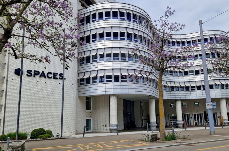 A modern, multi-story office building with a curved, circular design and large windows. The building has the word 'SPACES' prominently displayed on the facade. There are blooming trees in the foreground, and a taxi stand in front of the building.