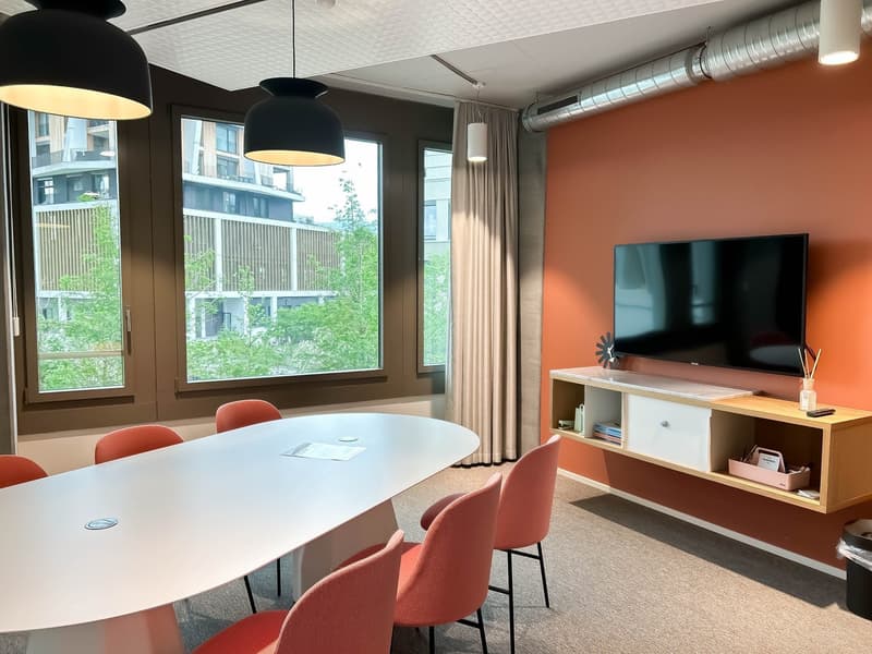 This image shows a modern and stylish office or meeting room space. The room features large windows that provide a view of the surrounding greenery outside. The room has a long white table with red chairs around it, as well as a TV mounted on the wall and