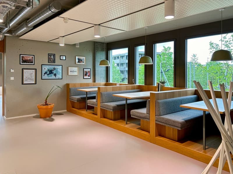 Modern dining area with wooden benches, tables, and pendant lighting. The space features large windows overlooking greenery outside. The walls are decorated with framed artwork and photographs.