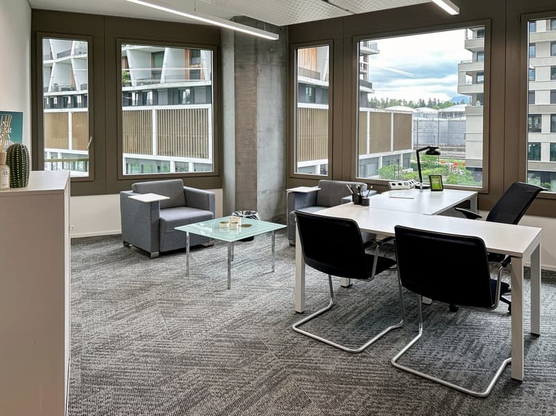 Modern office space with large windows, providing a view of the surrounding buildings. The room features a desk with a computer, chairs, a sofa, and a glass coffee table. The walls are dark-colored, and the floor is covered with gray carpeting.