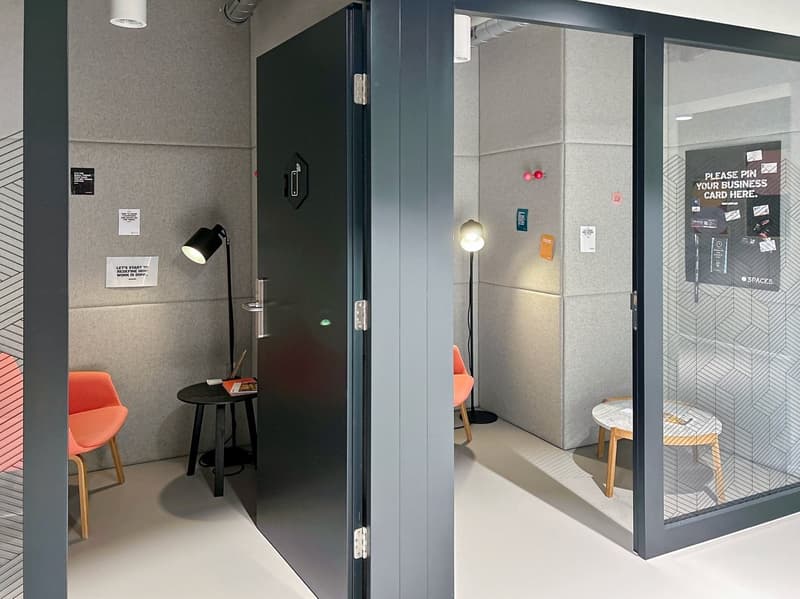 Meeting room with a black door, gray walls, small table, orange chairs, standing lamp, glass door.