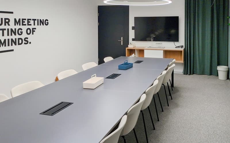 Long rectangular table, white chairs, black door, TV mounted on wall, wooden desk, green curtain, white trash bin