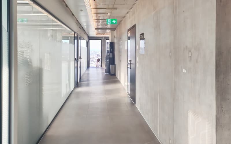 Long corridor with glass walls, tiled floor, emergency exit signs, and a trash bin.