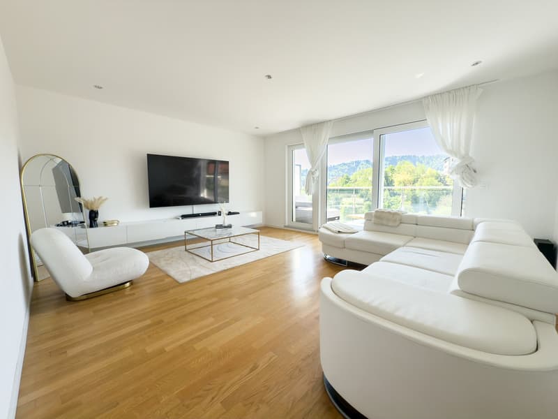 Spacious living room with hardwood floors, large windows providing a scenic view, white leather sofas, a TV mounted on the wall, and a gold-framed mirror on the side. The room has a modern and minimalist design aesthetic.