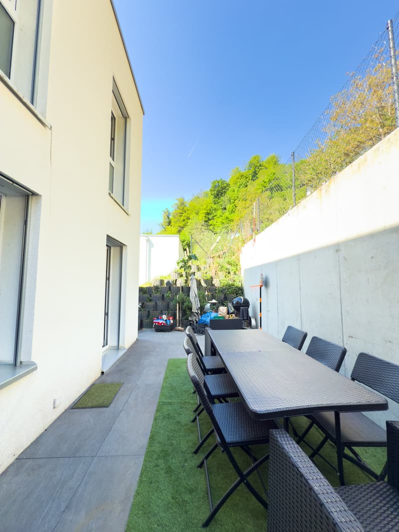 The image shows an outdoor patio area with a dining table and chairs, surrounded by lush greenery and a blue sky. The patio has a concrete floor and artificial grass, and there appears to be a balcony or terrace visible in the background.